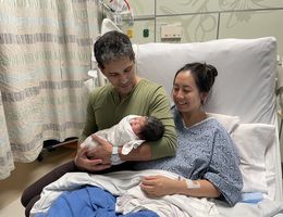 Married couple sits on bed together holding their newborn baby