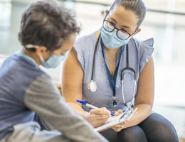 A Hispanic female doctor is taking down notes while speaking to a young 8 year old male patient. Both doctor and patient are wearing masks during the coronavirus pandemic.