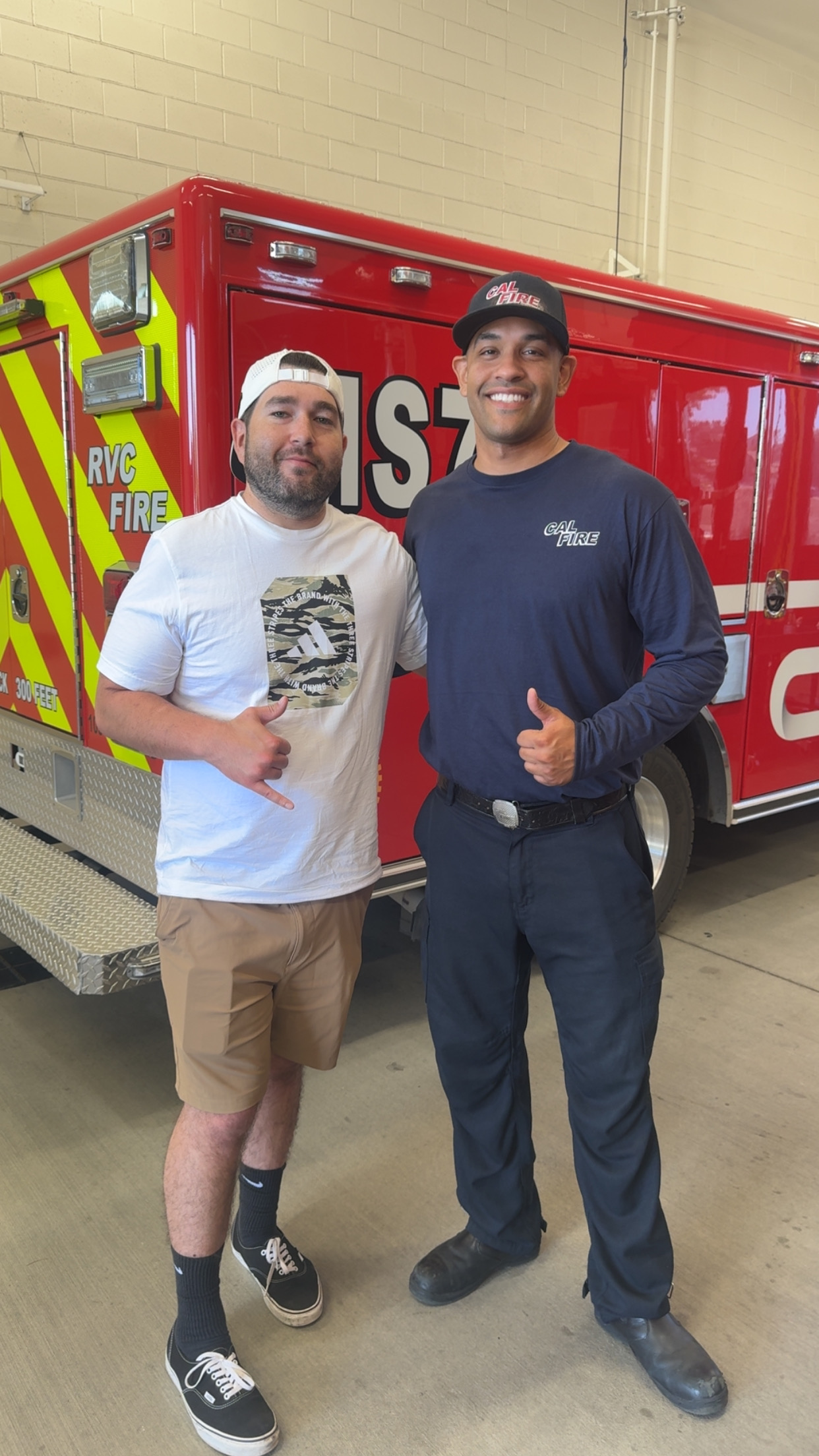 A young man stands next to a firefighter and both give the camera a thumbs up