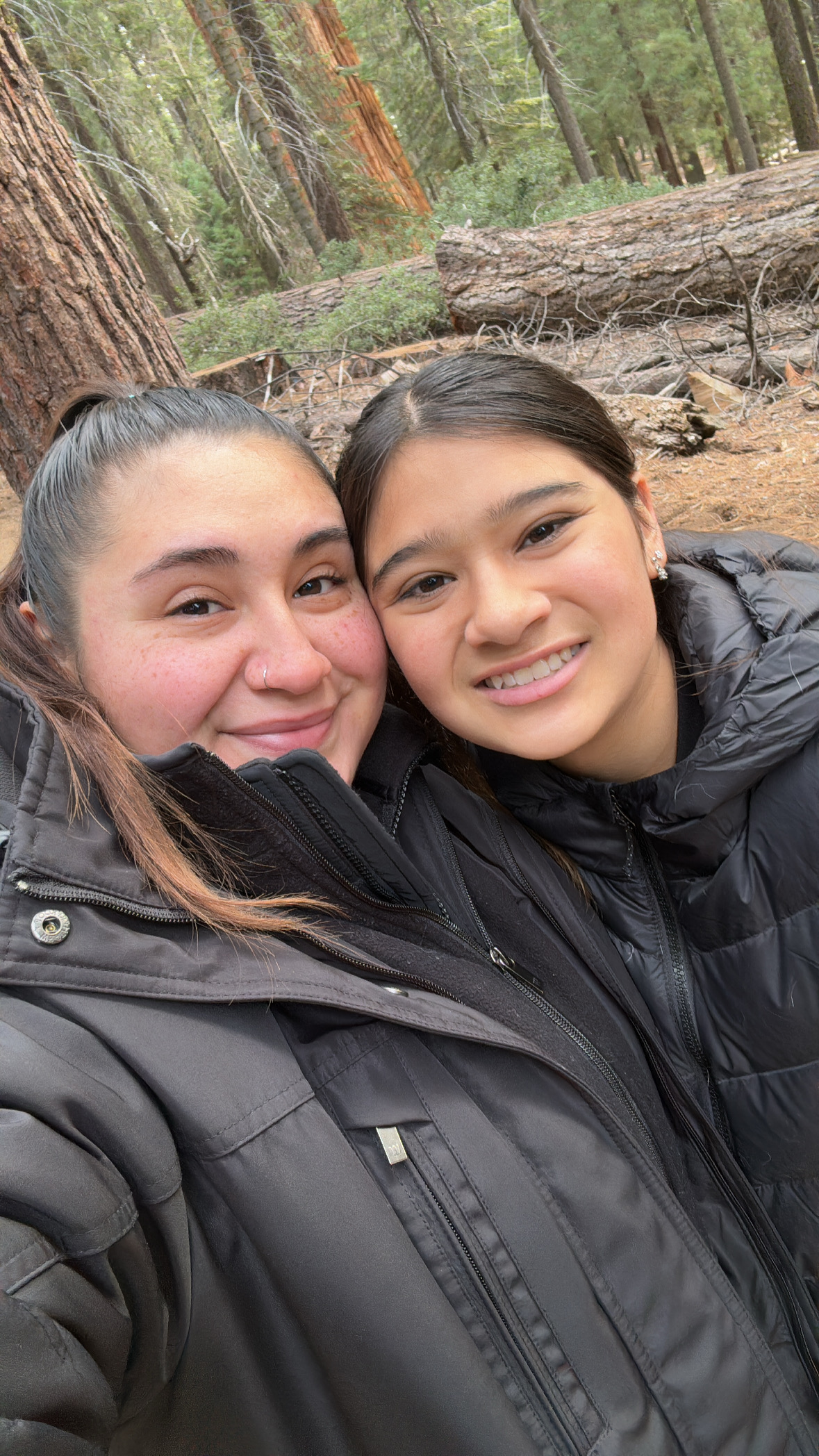 Two women in black jackets smile for a selfie in nature