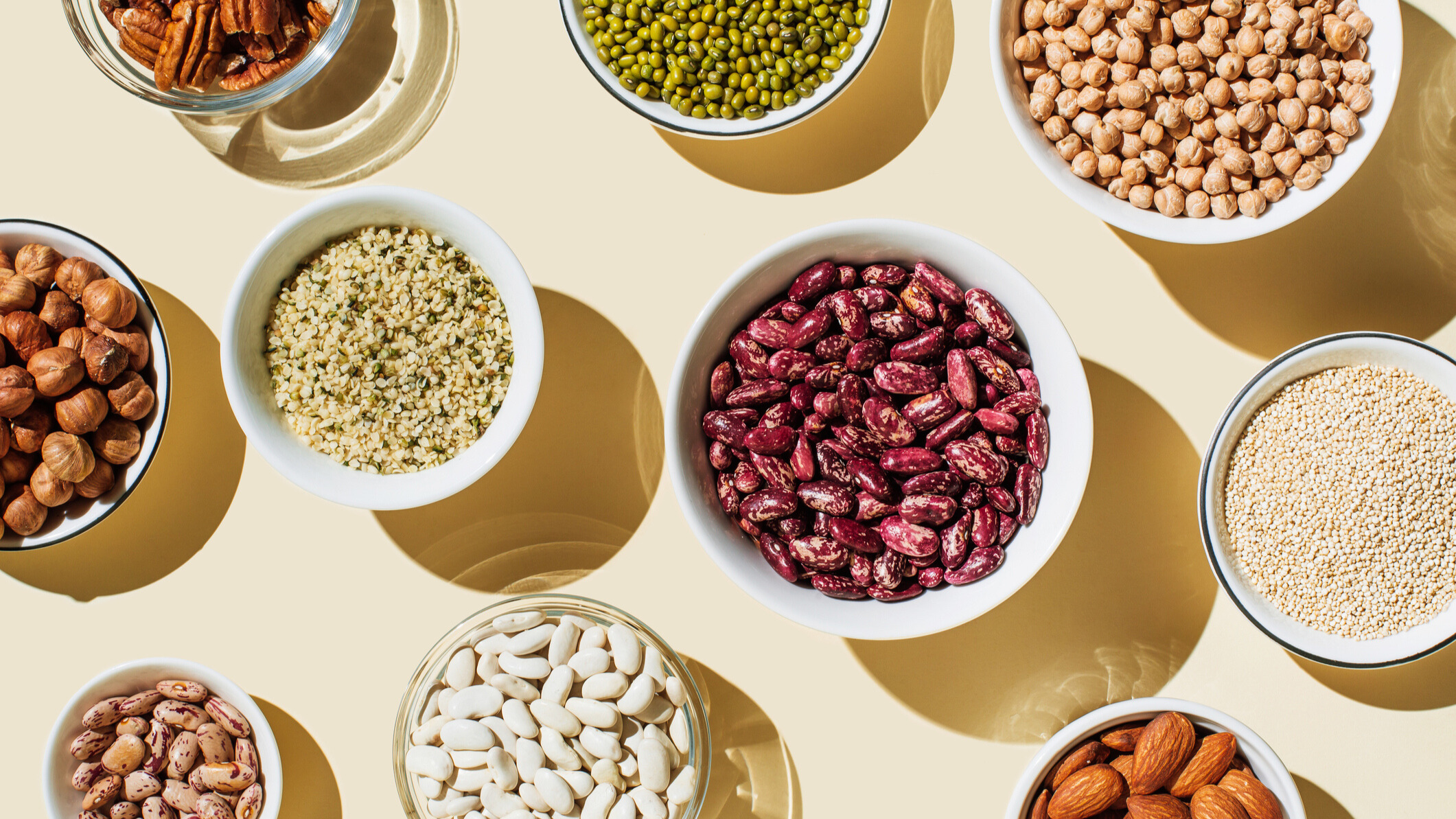 A top‑down view of various nuts, seeds, legumes, and grains arranged in small bowls on a light surface, including pecans, mung beans, chickpeas, hemp seeds, kidney beans, white beans, hazelnuts, almonds, and quinoa.