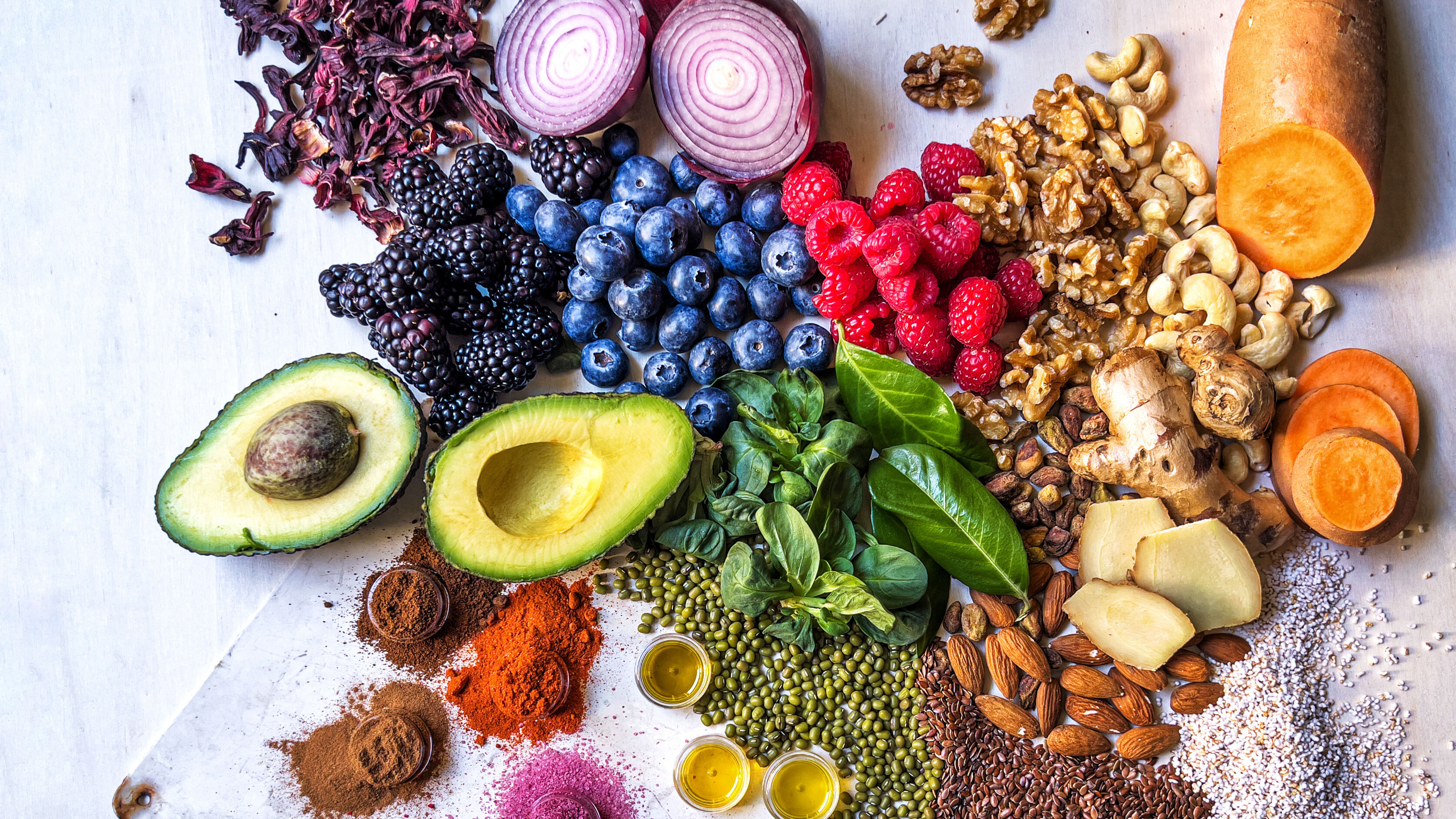 A colorful assortment of whole foods spread across a surface, including avocados, berries, leafy greens, sliced onions, sweet potatoes, nuts, seeds, spices, legumes, and small pools of oil, arranged in a vibrant flat‑lay style.