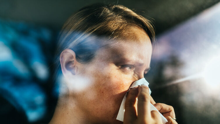 A person holding a tissue to their nose while sitting in a car, with light streaming through the window.