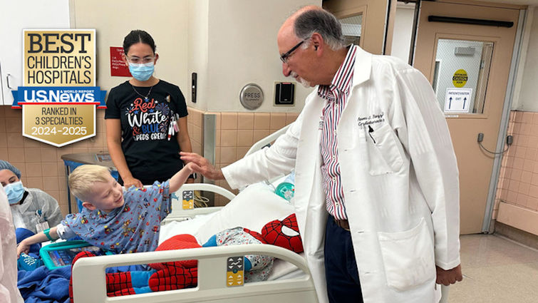 LLUCH patient Bryson Morris, 5, shares a high-five with his heart surgeon Anees Razzouk before undergoing a life-saving heart transplant on July 4 young blonde boy sits on hospital gurney in blue hospital gown giving his doctor wearing white coat and glasses a high five with two female medical staff nearby