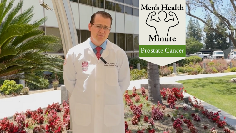 A doctor wearing a white coat stands with his hands behind his back while looking into the camera. He is in front of a medical building