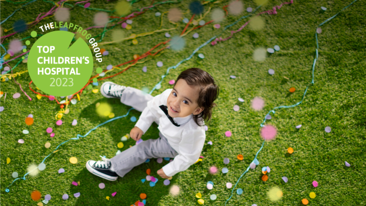 young boy with brown hair sitting in the grass, looking up with confetti falling.