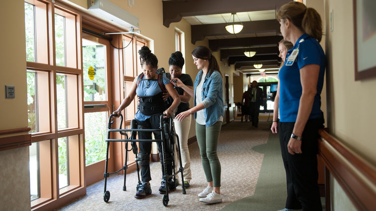Black woman uses an assisted walking device while receiving physical therapy with the help of two other women in a hallway next to a window.