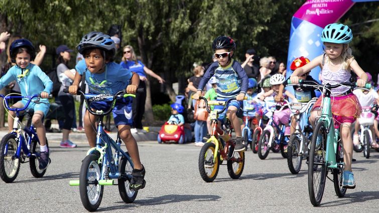 Bicycle riding can lead to traumatic brain injuries and concussions in children, with head injuries being the most common cause of serious disability and death in bicycle crashes. A group of young kids ride blue and yellow colored bikes all wearing helmets outside on the street at an event