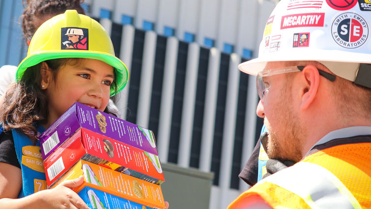 young girl scout holds colorful cookie boxes in front of kneeling construction worker