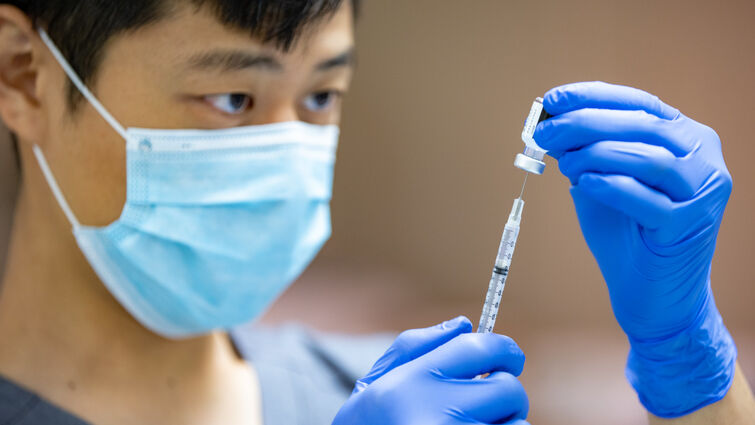 Asian male in blue scrubs, gloves, and surgical mask fills vaccine syringe.