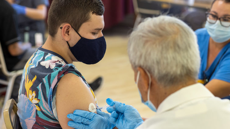 Doctor in white coat gives vaccine to person with short hair and colorful shirt