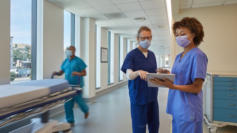 caucasion female nurse in navu scrubs and Black nurse in light blue scrubs in a hospital hallway, looking at a piece of paper