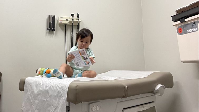 toddler sits on exam bed in clinic playing with toys and books before vaccine appointment