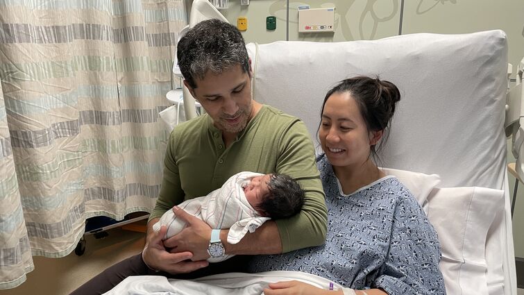 Married couple sits on bed together holding their newborn baby