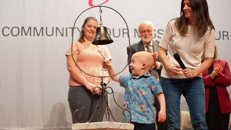 a young Caucasian boy with no hair in s blue shirt and jeans rings a standing bell, smiling with his mom beside him