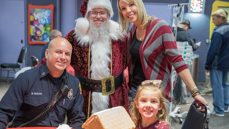 fireman, santa, mom, and kid pose for picture next to gingerbread house