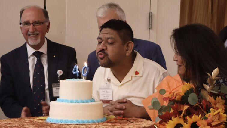 A man blows out candles on a two tiered cake with people surrounding him.