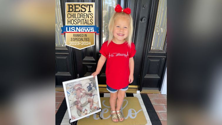 young blonde girl, smiling, holding a framed picture of herself as a baby undergoing treatment