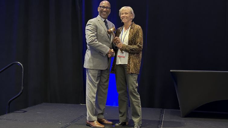 Craig Jackson takes the helm as President of the Association of Schools Advancing Health Professions, receiving the gavel from outgoing President Dr. Deb Larsen Black man in a grey business suit being presented an honorary gavel on stage by a Caucasion women in a pant suit,