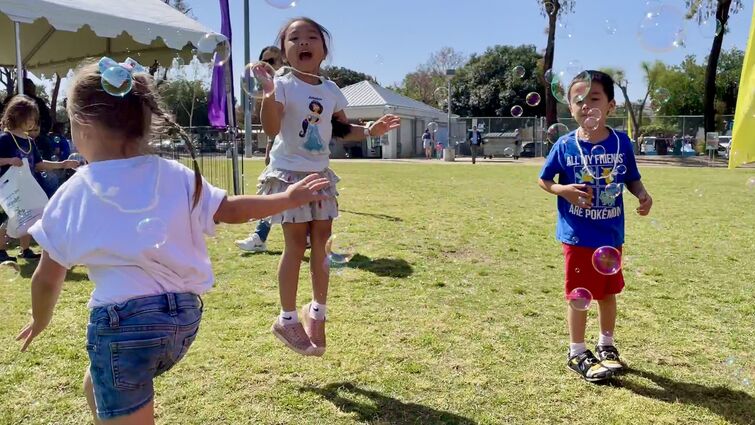 Three children in tee-shirts and shorts play outside in the grass blowing bubbles and smiling 