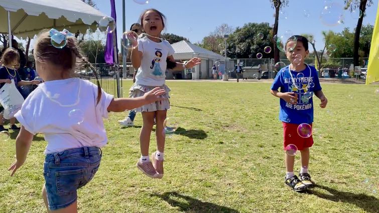 Three children in tee-shirts and shorts play outside in the grass blowing bubbles and smiling 