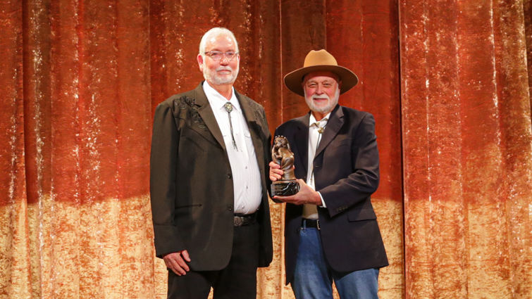 tall Caucasian man with white hair in a suit accepting award on stage from another Caucasian man in a suit and cowboy hat.