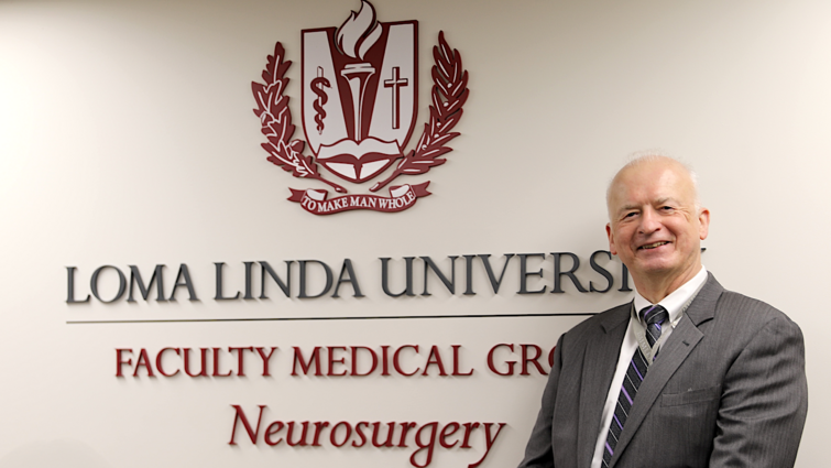 Man stands in front of a white wall with a sign saying Loma Linda University Faculty Medical Group Neurosurgery