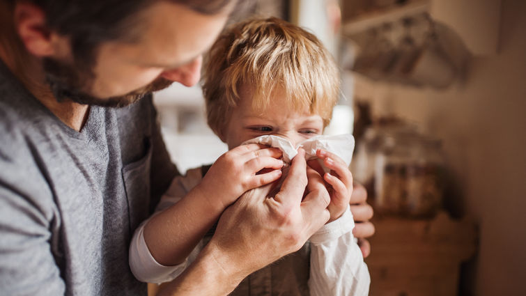 A father on paternity leave looking after small son indoors, blowing his nose