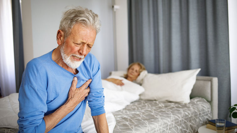 Senior man sitting on edge of bed holding chest in pain