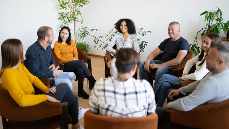 A group of mixed ethnic people sitting in a circle in comfortable chairs holding a support meeting, as they smile and chat with each other