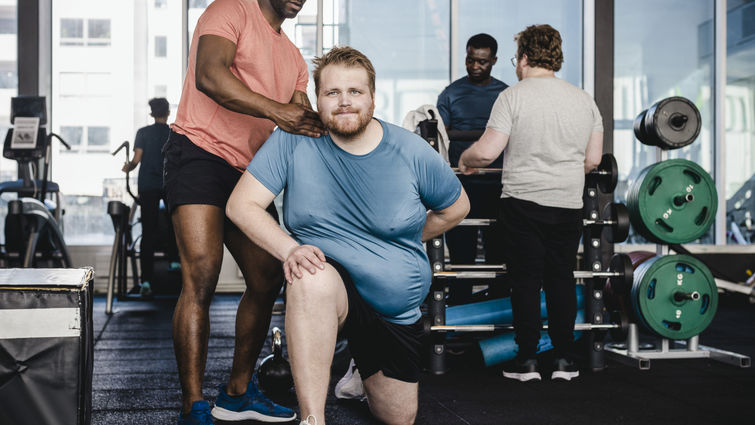 Male fitness instructor assisting overweight man in exercise at gym - stock photo