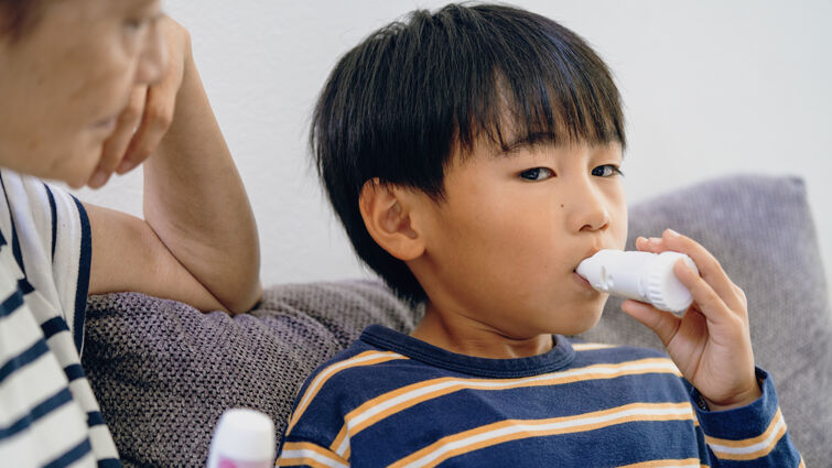 Boy using inhaler while sitting on gray couch, mom next to him supervising