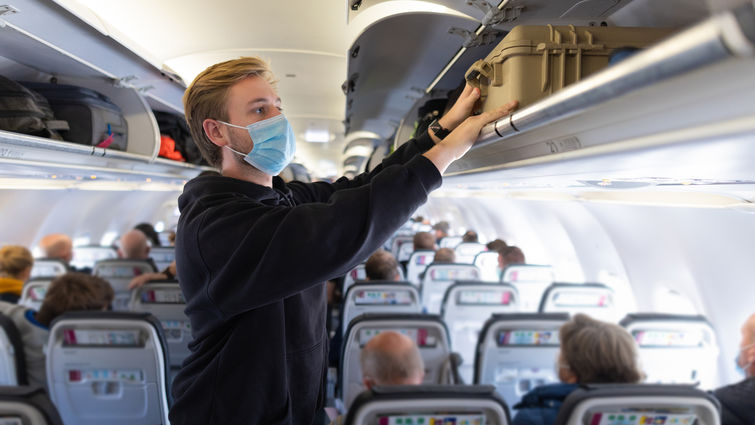 A young backpacker storing his luggage away while on a plane, budget travelling. He is wearing a protective face mask to reduce the spread of COVID-19.