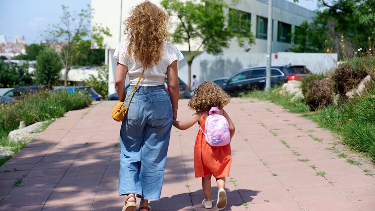 Parents can support their kids at every age to make the transition back to school a little easier Woman with curly hair holds hands with a child with curly hair and a backpack as they walk toward a school