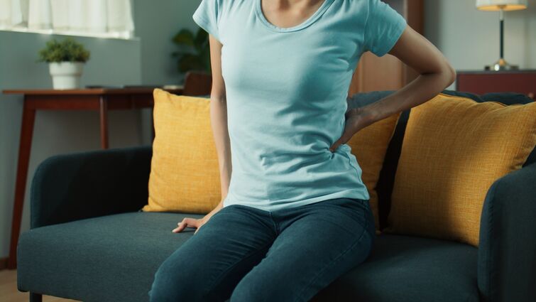 Persistent aches and pains could indicate something more serious Woman hand holding her low back, feeling low back pain and hurt while sits on sofa in living room at home