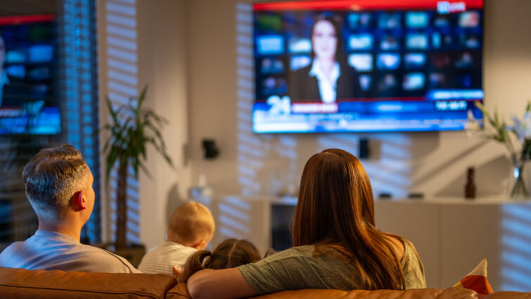 A woman, man, and baby sit on a brown couch and watch the news
