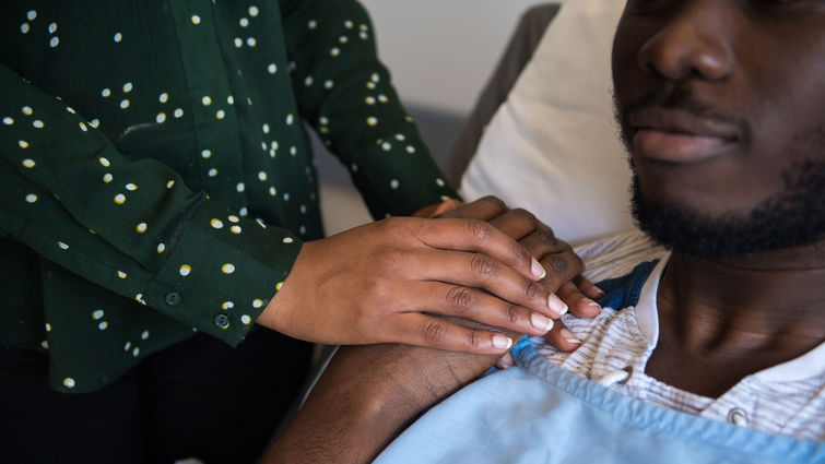 Woman holding man's hand as he lies in a hospital bed