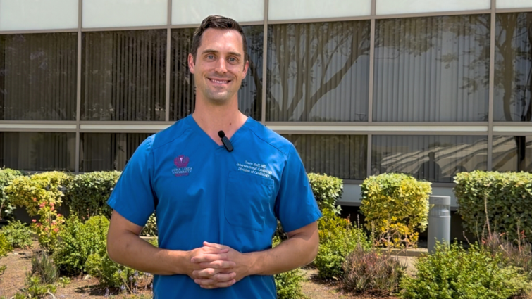 Doctor in blue scrubs stands outside a building surrounded by green bushes
