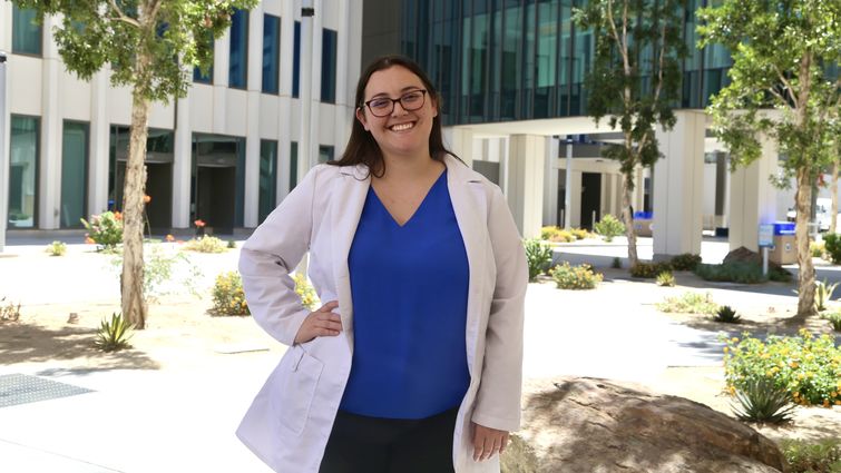 Caucasian woman standing outside in blue blouse and white coat with her right hand on her hip.