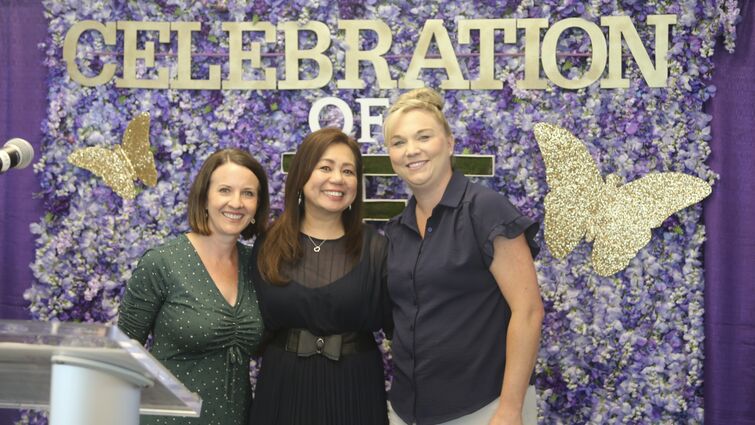 Three women stand smiling in front of purple flower backdrop with gold butterflies4. Sign reads "Celebration of Life"