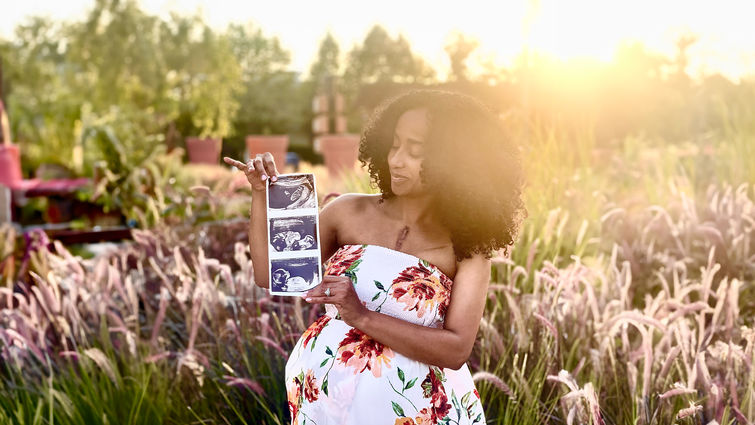 African American woman stands with sonogram in front of field, pregnant and smiling