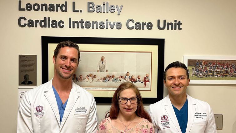 Colleen Barber reunites with the structural interventional cardiologists who recently performed a procedure to close a hole in her heart, Dr. Jason Hoff (left) and Dr. Amr Mohsen (right), framed by a background of photographs of Dr. Leonard Bailey who transplanted Barber's heart when she was a baby.