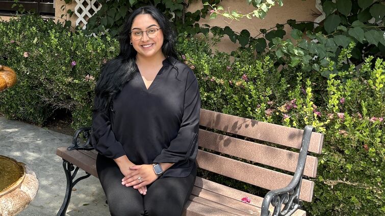 A woman in all black sits on a bench outside in front of beautiful flowers and greenary