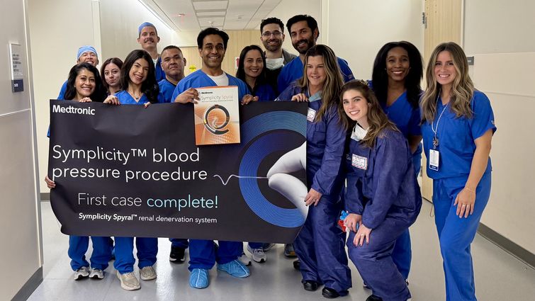 Vinoy Prasad, MD (fifth from left) Alec Kherlopian, MD, (fifth from right) and team celebrate the first renal denervation case in the region on March 11. Hospital staff all in blue scrubs stand smiling holding a banner that says "Symplicity blood pressure procedure. First case complete!"
