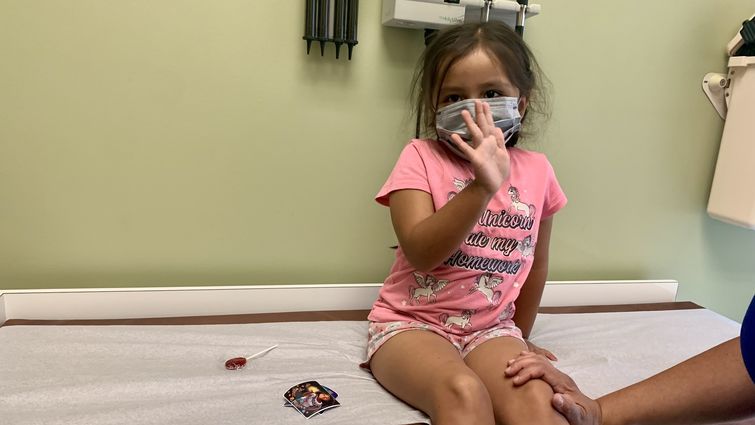 Three-year-old girl sits on exam table in clinic waving to camera about to get vaccination