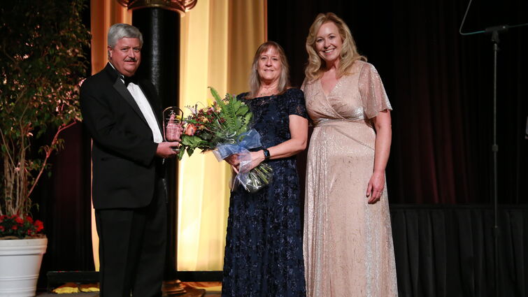 senior vice president for advancement, Rachelle, stands with Don and Penny holding award and flowers smiling for photo on stage