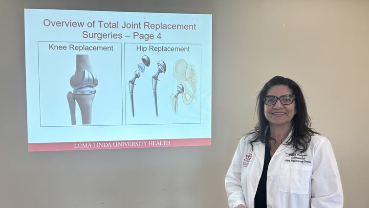 Female in a doctors white coat stands in front of a presentation slide on joint replacement