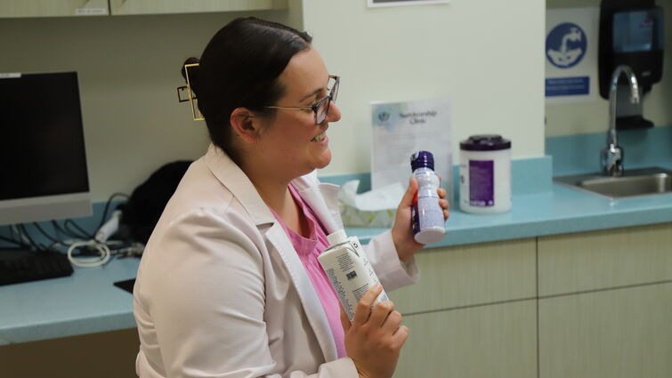 A woman is holding up two protein shakes as she talks to a patient in a medical room.