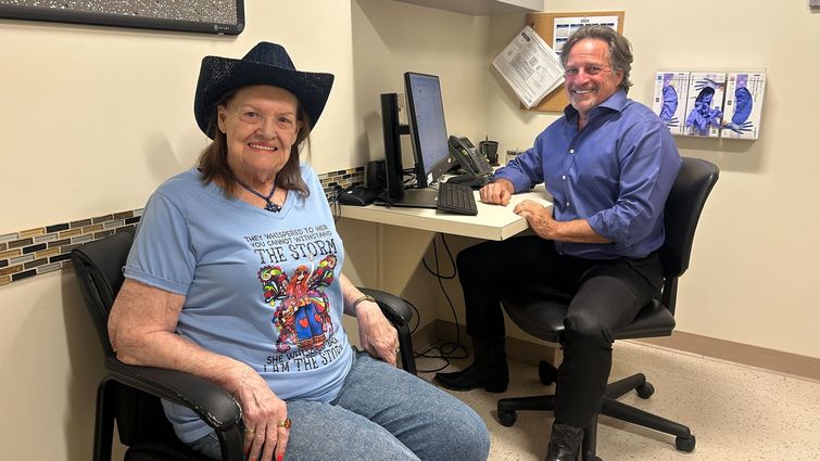 White woman sits smiling at camera in cowgirl cat next to middle aged white male physician. 