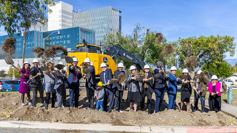 Hospital admin wearing hard hats stand in line in front of contruction vehicle and shoveling the ground 